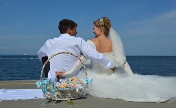 [Uber En="summer*wedding*couple*sitting*on*a*beach*looking*out*to*sea*with*a*picnic*basket*of*flowers*fruit*and*champagne" De="Sommerhochzeit*Paar*am*Strand*mit*Blick*aufs*Meer*neben*einem*Picknickkorb*mit*Blumen,*Obst*und*Sekt"]