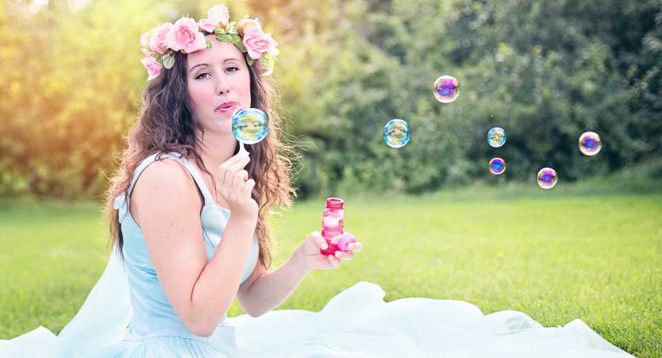 "Sommerhochzeit Braut mit rosanem Blumenkranz in einem hellblauen Kleid, die auf einer Wiese Seifenblasen bläst"